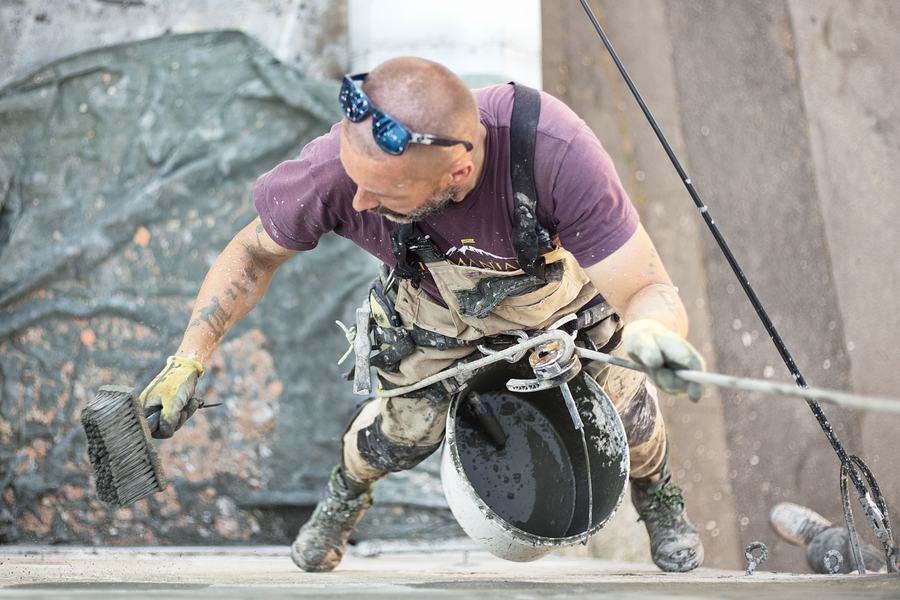 Peintre en baudrier suspendu devant un mur de façade avec un rouleau et un seau de peinture