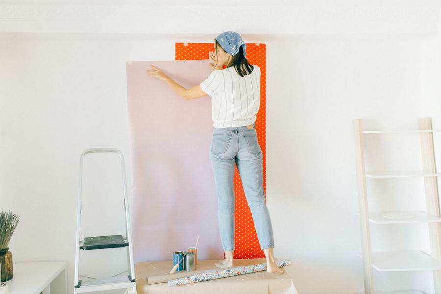 Femme sur un escabeau posant un lé de papier peint rose sur un mur blanc